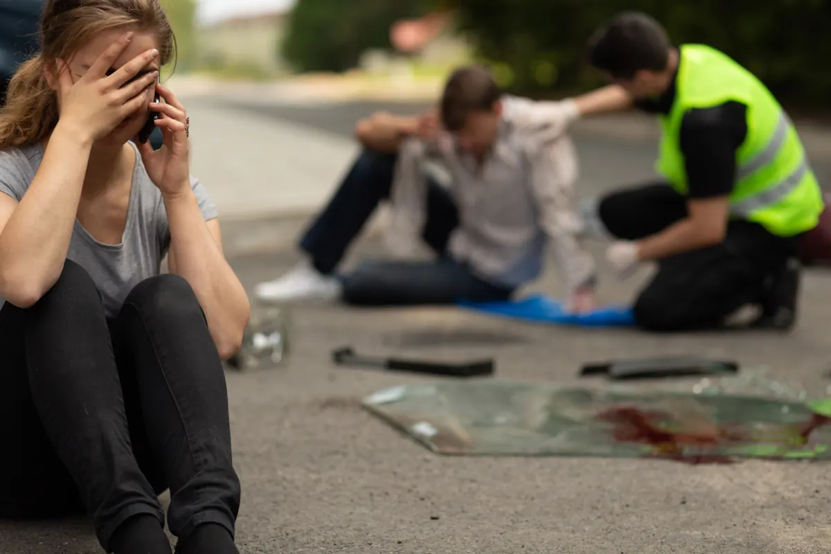 Es gibt viele Arten von möglichen Traumata. Ein schwerer Verkehrsunfall gehört dazu. Frau telefoniert mit den Händen im Gesicht auf der Straße sitzend, dahinter ein Helfer und ein verletzter Mann, auf dem Boden Spuren eines Verkehrsunfalls mit einem Auto